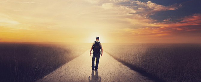 silhouette of man walking down a road towards sunrise