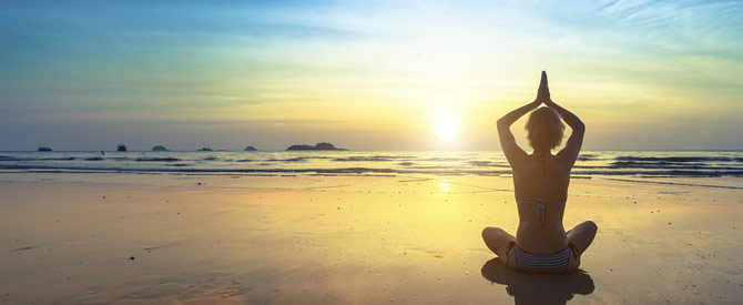 Woman sitting with hands clasped over her head in the prayer position. Silhouette while sitting on a beach at sunset.