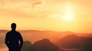Silhouette of man on hill looking out into the horizon at sunset pondering on life